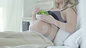 Unrecognizable pregnant woman lying on bed and eating healthful grapes. Relaxed Caucasian lady enjoying healthy food at