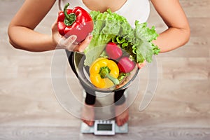 Unrecognizable rawfoodist girl checking weight on scale holding basket of fresh vegetables and green meat