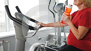 Unrecognizable senior caucasian woman drinking water from bottle during workout on training apparatus, looks cheerful