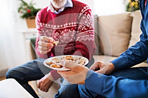 Unrecognizable senior father and adult son sitting on a sofa at Christmas time, eating biscuits.