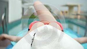 An unrecognizable slim busty woman in a big white hat and a pink bikini is lying on a chaise lounge near the pool. her