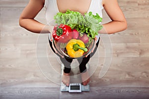 Unrecognizable slim girl checking weight on scale holding glass bowl with sweet pepper, fresh lettuce and redish