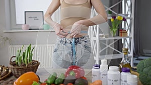 Unrecognizable slim woman measuring waist standing at table with healthful vegan food indoors. Young slender Caucasian