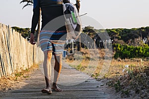 Unrecognizable surfer walks down the wooden walkway to the beach with a surfboard at sunrise