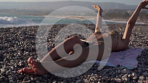 Unrecognizable tanned slim woman stretching sunbathing on pebble beach in sunset dusk. Relaxed happy Caucasian tourist