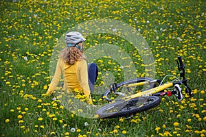 Unrecognizable teenage girl in a bicycle helmet sits with her back on the green grass among yellow dandelions