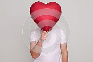 Unrecognizable unknown man holding red helium balloon in heart shape in front of his face guy wearing white casual T-shirt