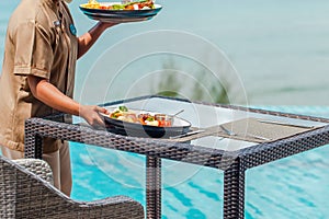 Unrecognizable waitress put food plates on table in outdoors restaurant at hotel