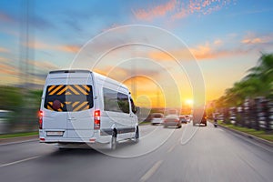 Unrecognizable white small passenger van hurry up on highway at city street traffic with urban cityscape, sunrise sky on