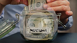 Unrecognizable woman add money to Saving Money In Glass Jar filled with Dollars banknotes. MORTGAGE transcription in