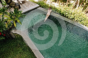 Unrecognizable woman in beige bikini relaxing in infinity pool in Bali admires a beautiful view of the palm trees.Luxury