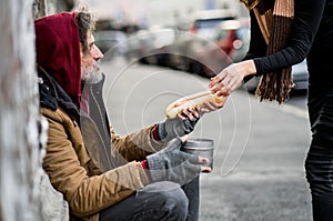 Unrecognizable woman giving food to homeless beggar man sitting in city.