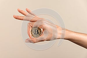 Unrecognizable woman holding bitcoin in manicured fingers of hand against beige backdrop