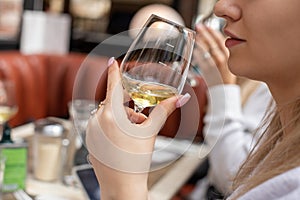 Unrecognizable woman holding clean glass with white wine near face. Adult lady in French restaurant tippling alcohol.