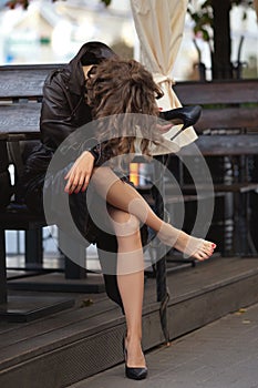 Unrecognizable woman in long coat sitting on bench and holding one shoe in her hand