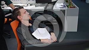 Unrecognizable woman passing waking up colleague sleeping on office chair. Portrait of tired overworking Caucasian young