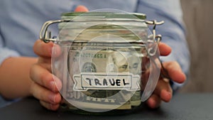 Unrecognizable woman put on table Saving Money In Glass Jar filled with Dollars banknotes. TRAVEL transcription in front