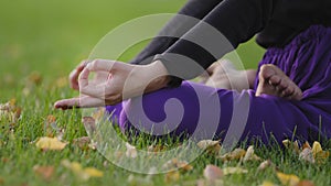 Unrecognizable woman sitting on grass lawn in park outdoors in lotus position with crossed legs doing yoga outside