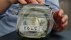 Unrecognizable woman turn Saving Money In Glass Jar filled with Dollars banknotes. 2025 year transcription in front of