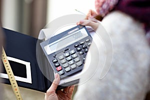 Unrecognizable woman in upper garments making calculations using calculator, measuring tape writing notes on notebook.