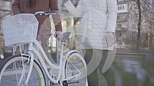 Unrecognizable woman in warm white jacket and tall man in brown coat walking on the street with a bicycle. Leisure of