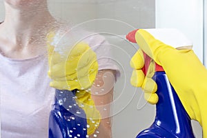 Unrecognizable woman in yellow rubber gloves holding a blue cleaning spray bottle and sprinkle on a glass mirror surface. Washing