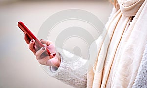 Unrecognizable young adult caucasian woman wearing jacket and scarf using her mobile smart phone outdoors on winter streets -