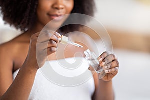 Unrecognizable young black woman in bath towel hlding bottle of face serum with pipette indoors, closeup