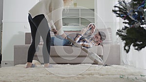 Unrecognizable young Caucasian woman vacuuming carpet at home when her husband lying on the couch. Wife throwing dirty