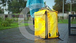 Unrecognizable young man in rain coat leaving yellow delivery backpack walking away in city park. Stressed Caucasian
