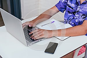 Unrecognizable young man working with laptop on terrace in swimsuit and blue hawaian shirt on white brick background.