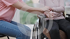 Unrecognizable young paralyzed woman and friend holding hands sitting indoors. Slim Caucasian empathic lady cheering up