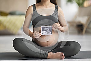 Unrecognizable young pregnant lady sitting on yoga mat and demonstrating baby sonography