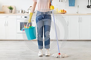 Unrecognizable young woman with bucket of cleaning supplies and mop ready to start wiping kitchen floor, indoors