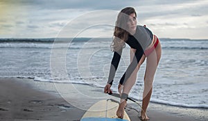 Unrecognizble barefoot woman has fixed legrope, stands on sand near surfboard, protects herself from crashing into shore lines,