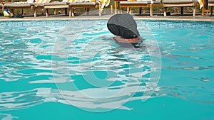 Unrecognized Caucasian senior elderly woman in black hat swims in blue water pool in hotel. Against the backdrop of