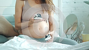 Unrecognized pregnant woman playing with baby shoes in bed.