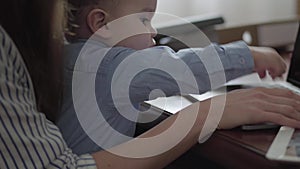 Unrecognized young mother is working at the table with laptop computer and documents folders. Small baby boy sit ay