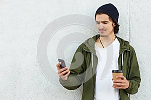 Unsatisfied disappointed youngster standing outdoors, spending time alone, checking social networks, holding his smartphone,