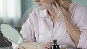 Unsatisfied woman in her 50s looking into a hand mirror, anti-age cosmetics