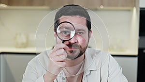 Unshaved brunette man looking at camera through magnifier and raising brow