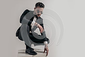 Unshaved young groom in black tuxedo crouching on wooden box