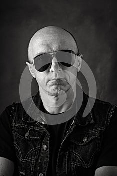 Unshaven, bald middle-aged man in a black T-shirt, denim vest and dark glasses. Black and white portrait