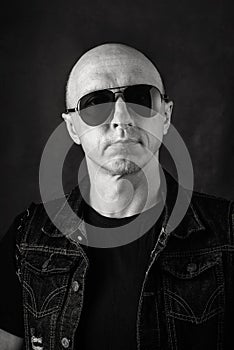 Unshaven, bald middle-aged man in a black T-shirt, denim vest and dark glasses. Black and white portrait