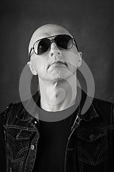 Unshaven, bald middle-aged man in a black T-shirt, denim vest and dark glasses. Black and white portrait