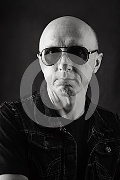 Unshaven, bald middle-aged man in a black T-shirt, denim vest and dark glasses. Black and white portrait