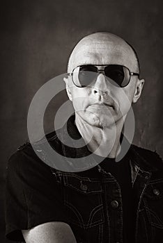 Unshaven, bald middle-aged man in a black T-shirt, denim vest and dark glasses. Black and white portrait toned in sepia