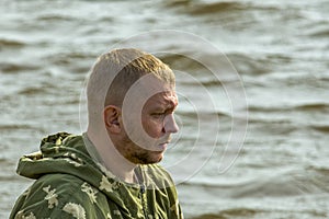 Unshaven fisherman against background of waves on river looks forward. Side view.