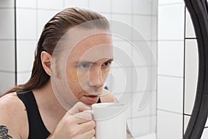 Unshaven man with patches under his eyes and cup of coffee looks at himself in bathroom mirror