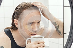 Unshaven man with patches under his eyes and cup of coffee looks at himself in the bathroom mirror. Male with morning hangover